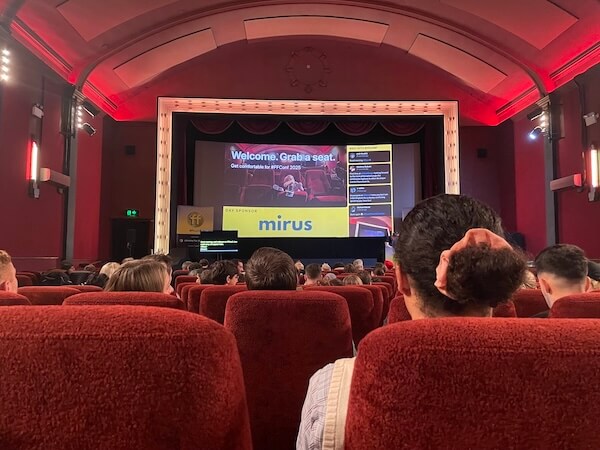 Theatre screen reads 'Welcome. Grab a seat. Get comfortable for ffconf 2025. Day sponsor: Mirus.' There's a sidebar with live posts from Bluesky. Audience sits in plushy red seats.